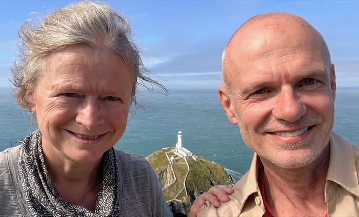 A close-up selfie of two people smiling with a lighthouse on a rocky outcrop in the background under a bright blue sky.
